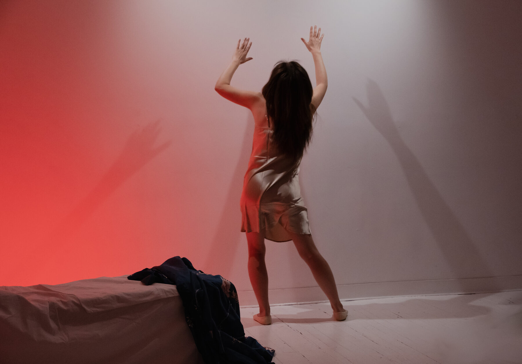A performer, lit in red, stands with arms raised against a bare studio wall, her shadow doubled behind her.